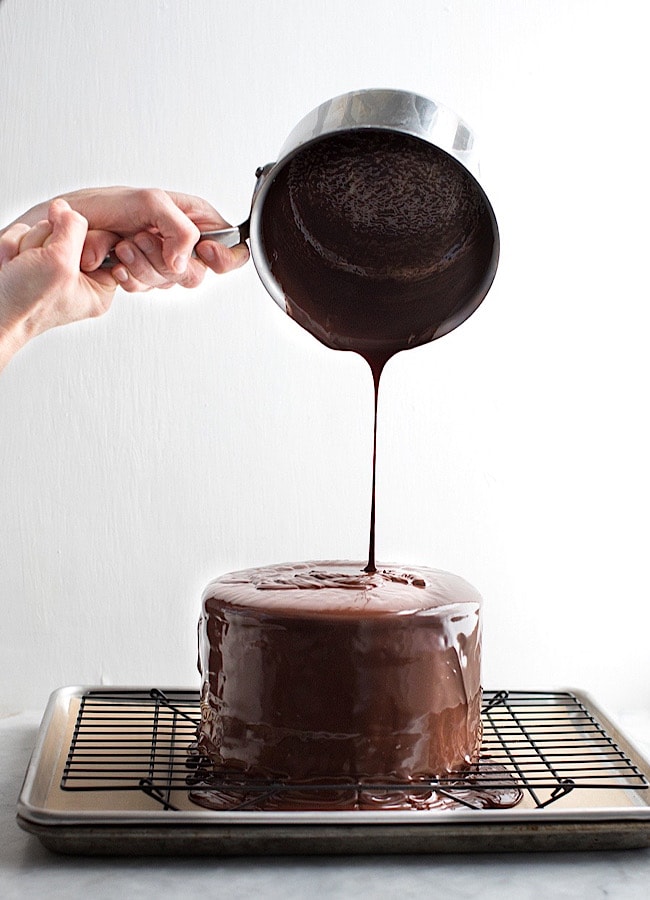 pouring chocolate ganache over a cake that is on a wire rack.